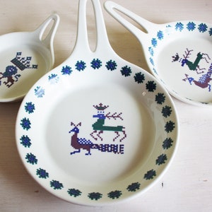 May include: Three white ceramic serving dishes with handles. Each dish features a different embroidered design in blue, green, and purple. The designs include a crown, deer, and bird. The dishes are on a white wooden surface.