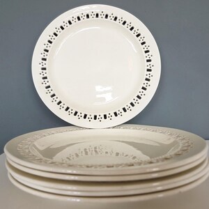 May include: A set of off-white ceramic plates with a decorative pierced border. One plate is positioned above the stack, highlighting the intricate design. The plates have a glossy finish and are set against a neutral backdrop.