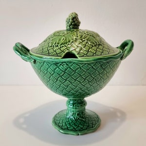 May include: A green ceramic serving bowl with a lid and two handles. The bowl is decorated with a woven pattern and has a grape cluster on top of the lid.
