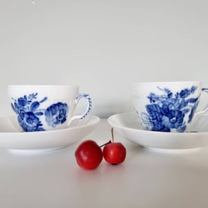 May include: Two white teacups with blue floral patterns and matching saucers. The cups and saucers have a textured surface. Two red apples are in the foreground.