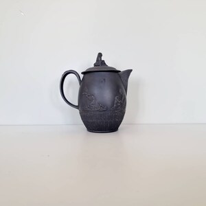 Op de afbeelding: Zwarte theepot van keramiek met een deksel en een handvat. De theepot heeft een reliëfdesign met figuren aan de zijkant.