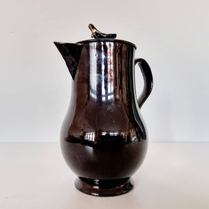 Puede incluir: Una jarra de cerámica negra con una tapa en forma de pájaro. La jarra tiene un cuerpo redondeado y un pico en un lado. La tapa está ligeramente entreabierta.