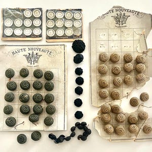 May include: A collection of vintage buttons including two small packs of white buttons labeled Frauengold, a large card of olive green textured buttons, a card of tan woven buttons, and a vertical row of black fabric buttons with a loose cluster.
