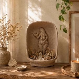 Può includere: Una scultura in pietra beige di forma ovale con un rilievo di una figura femminile che tiene in mano una luna crescente. La base della scultura contiene piccoli cristalli di quarzo trasparente e fiori secchi, creando un pezzo decorativo materico.