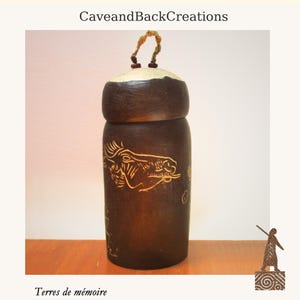 Puede incluir: Un recipiente cilíndrico de cerámica marrón oscuro con tapa y asa tejida. El recipiente presenta un diseño de cabeza de caballo grabado y el texto "CaveandBackCreations" y "Terres de mémoire".