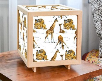 Op de afbeelding: Vierkante lamp met een licht houten frame, met een giraffenpatroon op de zijkanten. Het giraffendesign toont bruine en gele giraffen op een witte achtergrond. De lamp staat op kleine houten voetjes en is een decoratief verlichtingselement.