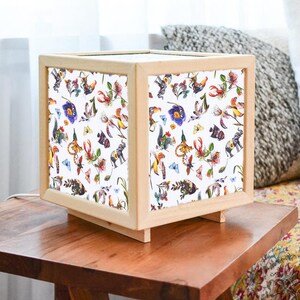Könnte beinhalten: Eine quadratische Holz-Lampenschirm mit weißem Hintergrund und bunten Tier- und Blumenillustrationen. Der Lampenschirm steht auf einem Holztisch.