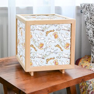 Könnte beinhalten: Ein Lampenschirm aus Holz in Würfelform mit weißem Hintergrund und einem Muster aus Eulen und Harry-Potter-Symbolen. Der Lampenschirm steht auf einem Holztisch.