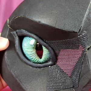 Puede incluir: Una máscara negra de forma geométrica con un ojo grande y detallado. El ojo es una vibrante mezcla de verde y azul, con una malla que cubre parte del ojo. La máscara está hecha de un material texturizado.