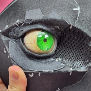 Puede incluir: Primer plano de una máscara negra con un ojo realista. El ojo es de un verde vibrante con iris y venas detalladas. La máscara tiene una sección de malla y está sujeta por una mano.