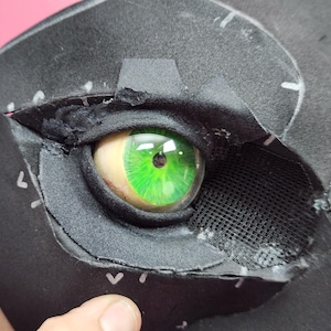 Puede incluir: Primer plano de una máscara negra con un ojo verde vibrante. El ojo tiene una apariencia realista con un iris y una pupila detallados. La máscara está construida con materiales en capas, con costuras visibles y detalles de malla alrededor del ojo.