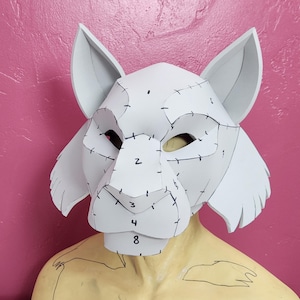 Pode incluir: Máscara de espuma branca com seções numeradas, que se assemelha a uma cabeça de lobo ou raposa. A máscara está no torso de um manequim.