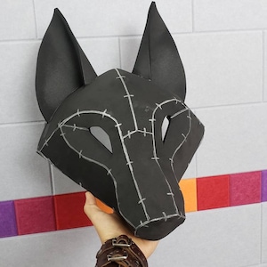 Puede incluir: Una m&aacute;scara de lobo negra con orejas puntiagudas y aberturas para los ojos. La superficie de la m&aacute;scara est&aacute; construida con m&uacute;ltiples paneles unidos por l&iacute;neas de costura de color gris claro que crean un patr&oacute;n geom&eacute;trico en la cara.