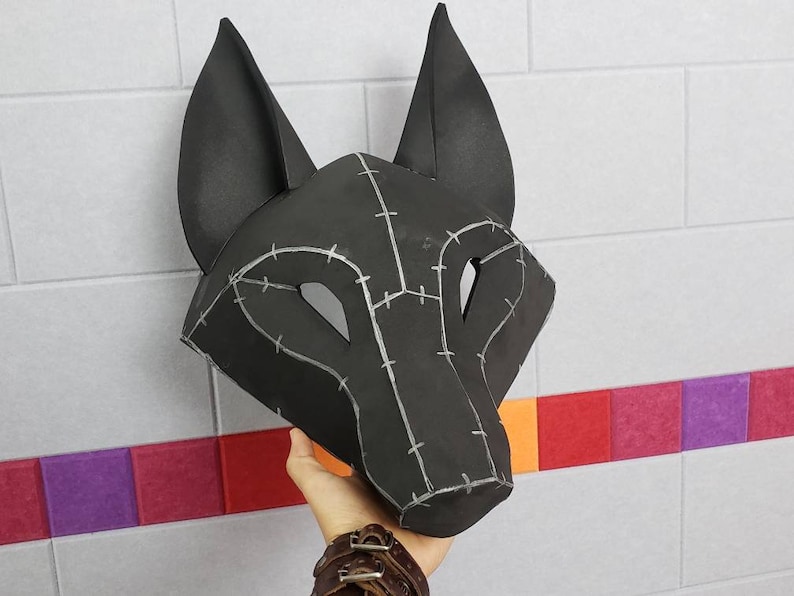 Puede incluir: Una m&aacute;scara de lobo negra con orejas puntiagudas y aberturas para los ojos. La superficie de la m&aacute;scara est&aacute; construida con m&uacute;ltiples paneles unidos por l&iacute;neas de costura de color gris claro que crean un patr&oacute;n geom&eacute;trico en la cara.