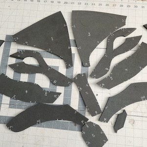 May include: A set of 12 black foam pieces, cut into various shapes and sizes, with numbers and letters marked on them. The pieces are laid out on a white gridded surface.