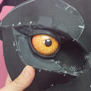 Puede incluir: Primer plano de una máscara negra con un ojo ámbar detallado. El ojo presenta vetas intrincadas y una pupila negra. Una malla cubre la parte inferior del ojo. La máscara está hecha de un material oscuro con costuras y marcas visibles.