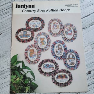 May include: A leaflet for cross-stitch patterns featuring country rose ruffled hoops. The leaflet shows 10 different designs with text such as "Welcome", "The kitchen is the heart of our home", and "Bless my little kitchen with love".