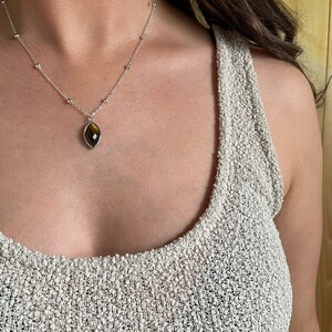 May include: Silver necklace with a marquise-shaped pendant featuring a brown and gold gemstone. The necklace has small silver beads along the chain. The person is wearing a light beige knit top.