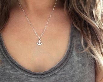 Puede incluir: Un collar plateado con un pequeño colgante de ancla. El ancla cuelga de una fina cadena. El collar se lleva sobre una camiseta sin mangas gris. La imagen es un primer plano del collar y la parte superior del pecho.