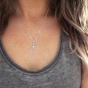 May include: A silver-colored necklace with a small anchor pendant. The anchor is centered on a delicate chain. The necklace is worn over a gray tank top. The image is a close-up of the necklace and upper chest.