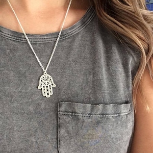 Könnte beinhalten: Eine silberne Halskette mit einem zarten, filigranen Hamsa-Hand-Anhänger. Die Kette wird von einer Person getragen, die ein graues T-Shirt mit einer Tasche trägt.