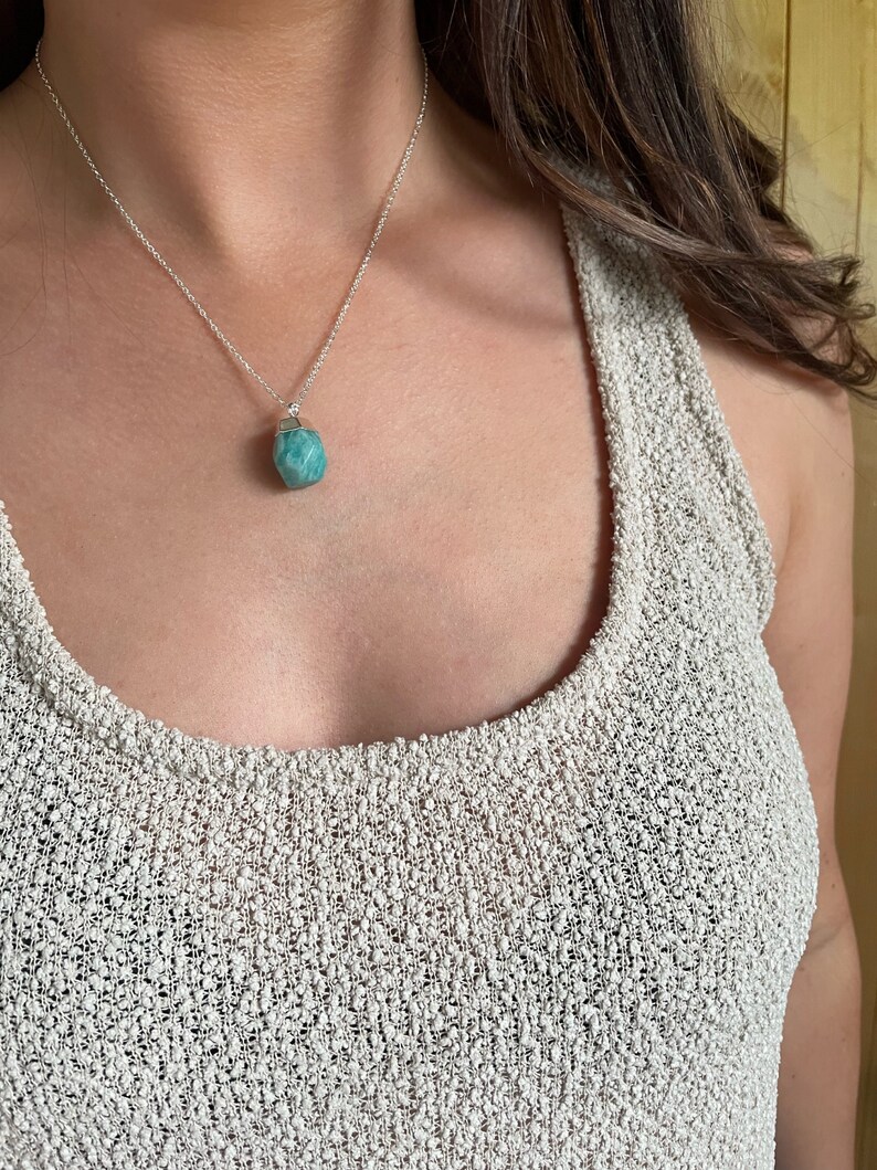 May include: A silver necklace with a small, light blue gemstone pendant. The pendant is a rough, irregular shape. The necklace is worn over a light-colored, textured top.