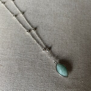Op de afbeelding: Een zilveren ketting met een hanger. De hanger heeft een ovale vorm met een lichtgroene steen. De fijne ketting heeft kleine zilveren kralen. De ketting ligt op een neutraal gekleurde stof.