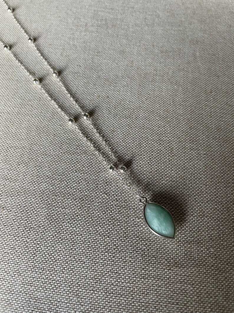 Op de afbeelding: Een zilveren ketting met een hanger. De hanger heeft een ovale vorm met een lichtgroene steen. De fijne ketting heeft kleine zilveren kralen. De ketting ligt op een neutraal gekleurde stof.