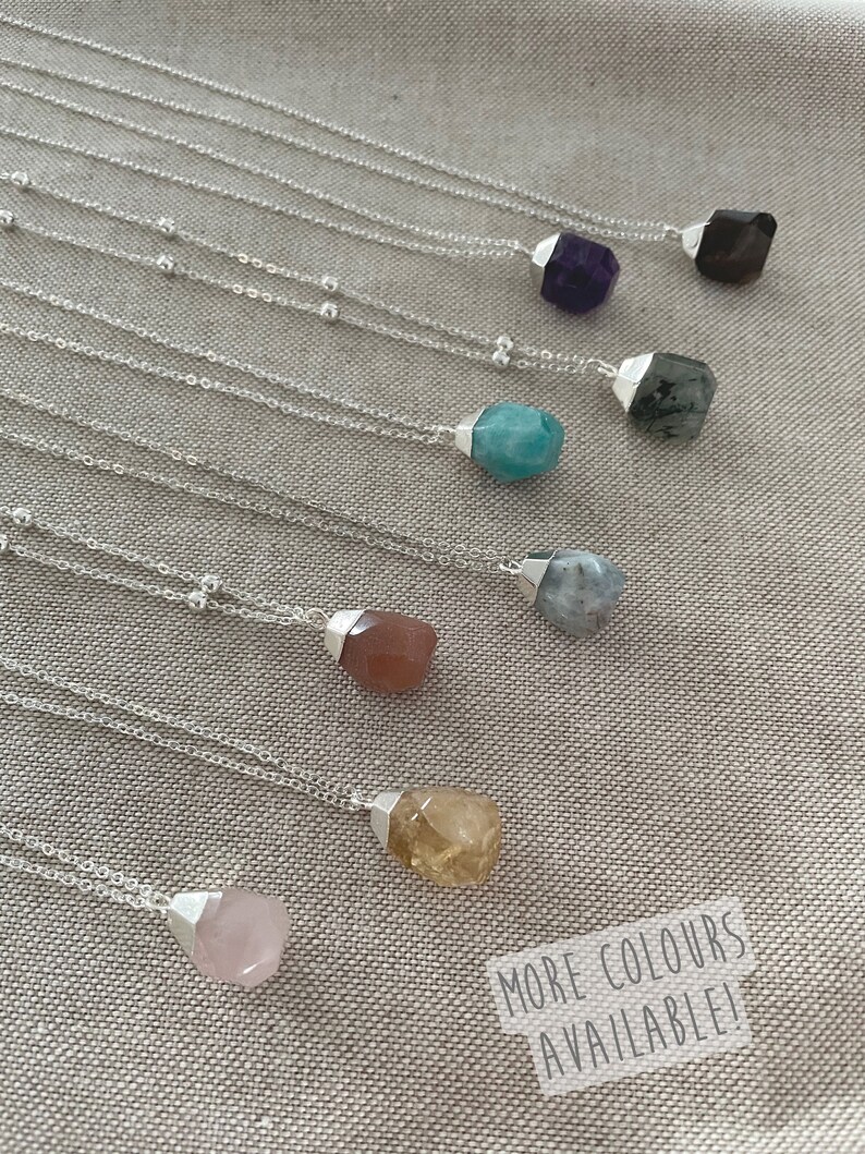 May include: A collection of gemstone necklaces with silver chains. The gemstones are various colors, including purple, blue, brown, yellow, and pink. The text "MORE COLOURS AVAILABLE!" is visible in the bottom right corner.