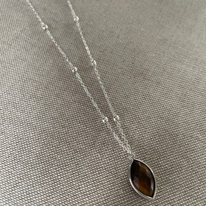 May include: A silver necklace with a brown, marquise-shaped pendant. The pendant is set in a silver bezel. The necklace chain features small silver beads and a delicate chain. The jewelry is displayed on a textured, neutral-colored surface.