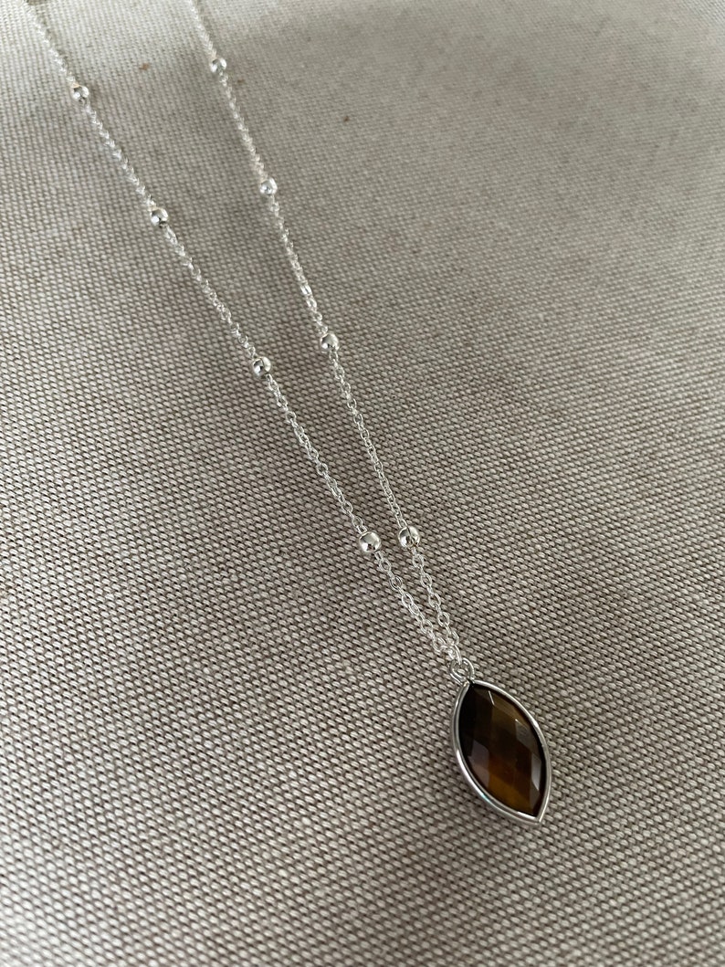 May include: A silver necklace with a brown, marquise-shaped pendant. The pendant is set in a silver bezel. The necklace chain features small silver beads and a delicate chain. The jewelry is displayed on a textured, neutral-colored surface.