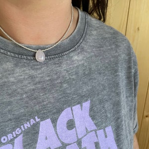 May include: A silver chain necklace with a light pink teardrop-shaped pendant. The necklace is worn by a person wearing a gray t-shirt with the text "ORIGINAL BLACK" in purple.