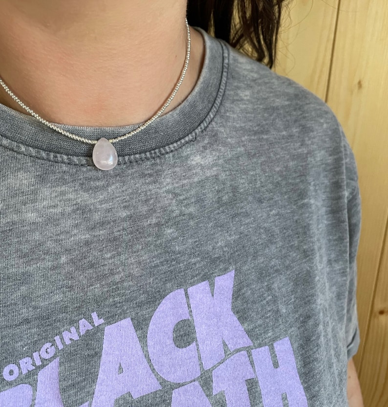 May include: A silver chain necklace with a light pink teardrop-shaped pendant. The necklace is worn by a person wearing a gray t-shirt with the text "ORIGINAL BLACK" in purple.