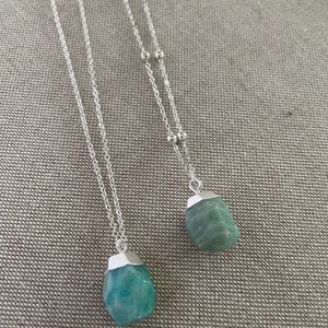 May include: Two silver necklaces with pendants. Each necklace features a silver chain with a small, irregular, light blue-green stone pendant. The stones have a silver cap at the top. The necklaces are displayed on a neutral-colored surface.