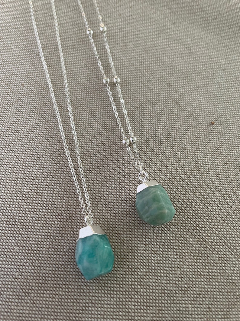 May include: Two silver necklaces with pendants. Each necklace features a silver chain with a small, irregular, light blue-green stone pendant. The stones have a silver cap at the top. The necklaces are displayed on a neutral-colored surface.