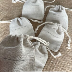 May include: A collection of six small, beige drawstring bags made of a textured fabric. Each bag features a simple arrow design and a drawstring closure. The bags are arranged on a textured, neutral-toned surface. The text "ECO-FRIENDLY PACKAGING!" is visible.