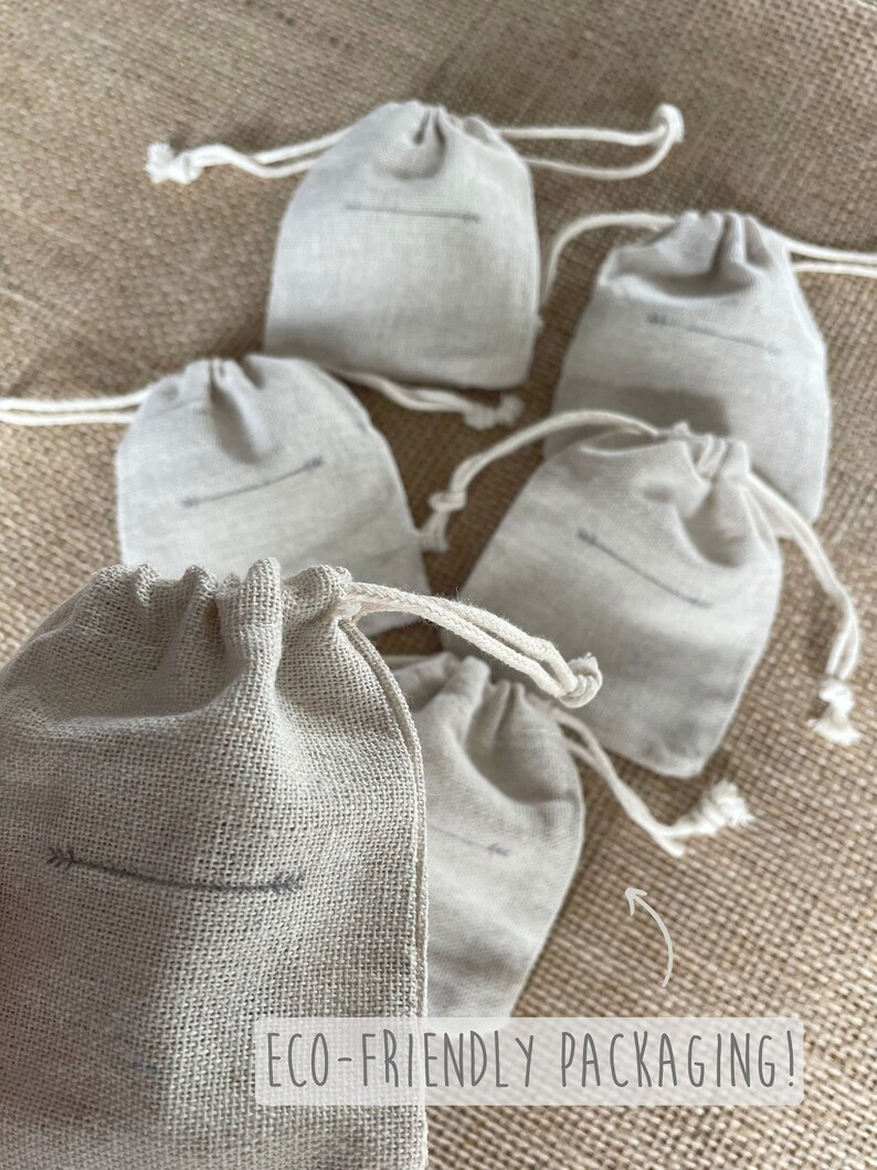 May include: A collection of six small, beige drawstring bags made of a textured fabric. Each bag features a simple arrow design and a drawstring closure. The bags are arranged on a textured, neutral-toned surface. The text "ECO-FRIENDLY PACKAGING!" is visible.