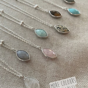 May include: A collection of silver necklaces with teardrop-shaped gemstone pendants in various colors, including gray, pink, and turquoise. The necklaces are displayed on a neutral-colored fabric. The text "MORE COLORS AVAILABLE!" is visible.