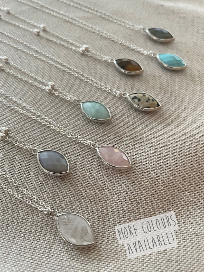 May include: A collection of silver necklaces with teardrop-shaped gemstone pendants in various colors, including gray, pink, and turquoise. The necklaces are displayed on a neutral-colored fabric. The text "MORE COLORS AVAILABLE!" is visible.