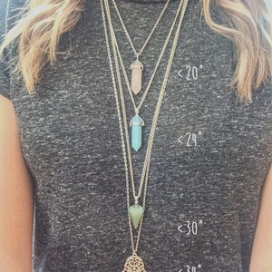 May include: Four silver necklaces with different length pendants. The shortest necklace has a light pink crystal pendant, the next has a light blue crystal pendant, the third has a green triangle pendant, and the longest has a silver hamsa hand pendant. The necklaces are labeled with their lengths: <20", <24", <30", and <34".