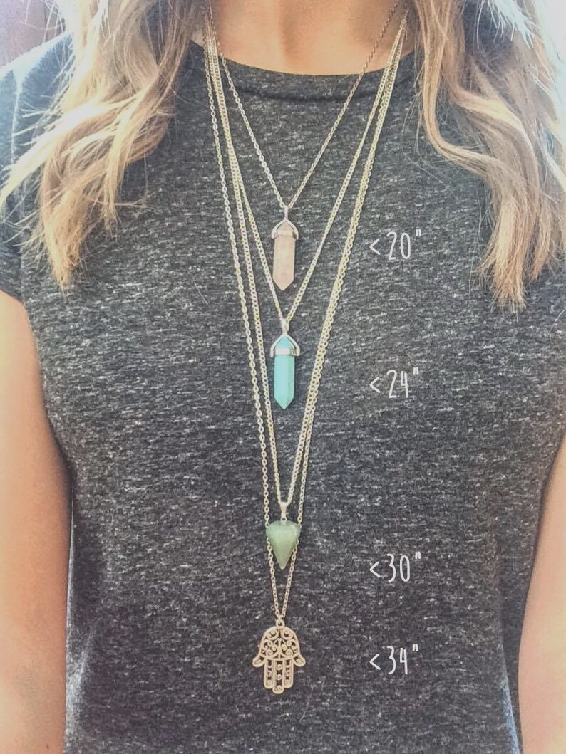 May include: Four silver necklaces with different length pendants. The shortest necklace has a light pink crystal pendant, the next has a light blue crystal pendant, the third has a green triangle pendant, and the longest has a silver hamsa hand pendant. The necklaces are labeled with their lengths: <20", <24", <30", and <34".