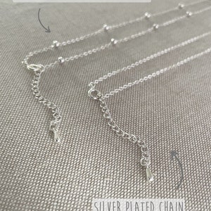 May include: Close-up of two silver-plated necklaces. One is a satellite chain with small, evenly spaced beads. The other is a silver-plated chain. Both have adjustable clasps and are displayed on a textured, neutral-toned surface.