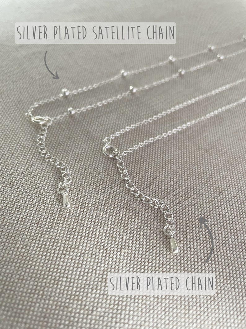 May include: Close-up of two silver-plated necklaces. One is a satellite chain with small, evenly spaced beads. The other is a silver-plated chain. Both have adjustable clasps and are displayed on a textured, neutral-toned surface.