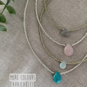 May include: Four silver beaded necklaces with different colored gemstone pendants. The pendants are turquoise, light blue, pink, and gray. The necklaces are layered on a beige fabric background. The text "MORE COLORS AVAILABLE!" is printed on a white label.
