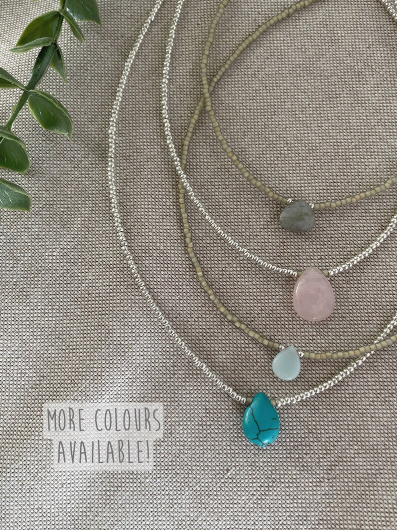 May include: Four silver beaded necklaces with different colored gemstone pendants. The pendants are turquoise, light blue, pink, and gray. The necklaces are layered on a beige fabric background. The text "MORE COLORS AVAILABLE!" is printed on a white label.