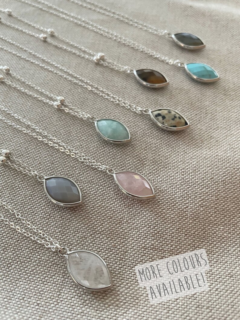 Op de afbeelding: Een verzameling zilveren kettingen met druppelvormige edelsteenhangers in diverse kleuren, waaronder grijs, roze en turquoise. De kettingen zijn gepresenteerd op een neutrale stof. De tekst "MORE COLOURS AVAILABLE!" is zichtbaar.