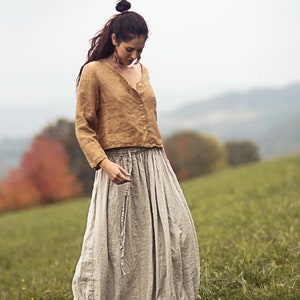 Può includere: Una donna indossa una blusa di lino marrone e una lunga gonna di lino grigio chiaro. È in piedi in un campo di erba verde.