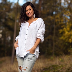 May include: A woman wearing a white linen shirt with a button-down front and blue jeans with distressed details. She is standing in a forest setting with blurred trees in the background.