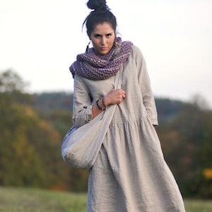 Può includere: Una donna indossa un abito di lino beige chiaro con una sciarpa di lana viola e una grande borsa tote. È in piedi in un campo di erba verde.