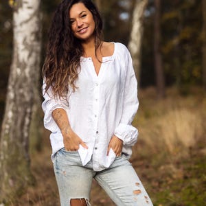 May include: A woman with long, wavy brown hair is wearing a white linen shirt and light blue jeans with distressed details. She is standing in a forest setting with trees in the background.